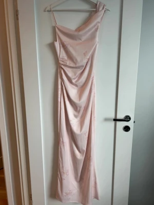 Adoore klänning  - Elegant ljusrosa långklänning i satin med oneshoulder-design och smala axelband. Klänningen har draperad front och subtilt blommigt mönster som ger en romantisk känsla. Perfekt för speciella tillfällen när du vill känna dig extra fin.