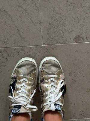 Golden Goose Superstar sneakers - Golden Goose Superstar sneakers i beige och guld med ormmönstrade detaljer, svart stjärna på sidan och blå hälflik. Skorna har vita skosnören och vit sula. Klassisk low-top modell med cool vintagekänsla och unika materialmixar.