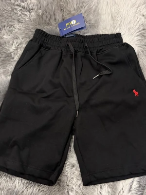 Svarta shorts från Polo Ralph Lauren - Svarta shorts från Polo Ralph Lauren med elastisk midja och snörning. Klassisk röd broderad logga på vänster ben och två sidofickor. Tillverkade i mjuk bomull, perfekta för chill dagar eller när du vill ha en clean look.