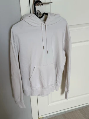 Vit hoodie med huva och magficka - Klassisk vit hoodie med huva och dragsnören samt en stor magficka framtill. Hoodien har långa ärmar och ribbade muddar vid ärmslut och nederkant. Perfekt för en avslappnad och stilren look.