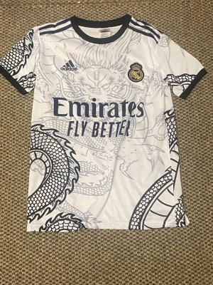 Real Madrid draktröja Adidas vit - Säljer en vit Real Madrid matchtröja från Adidas med svart drakmönster över hela tröjan. Tröjan har svarta detaljer vid krage och ärmslut, samt klubbmärke och Adidas-logga på bröstet. Tillverkad i lätt och ventilerande material, perfekt för fotbollsträning.