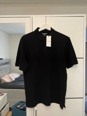 Svart pikétröja från Jack & Jones - Klassisk svart pikétröja från Jack & Jones med korta ärmar och krage. Tröjan har diskret logga broderad på bröstet och stängs med knappar framtill. Perfekt till både jeans och shorts för en clean look.