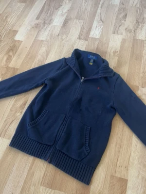 Mörkblå ziptröja från Polo Ralph Lauren - Mörkblå ziptröja från Polo Ralph Lauren med ribbade muddar, hög krage och två framfickor. Tröjan har en liten röd logga broderad på bröstet och är tillverkad i mjukt material. Perfekt för lager på lager och enkel att matcha.