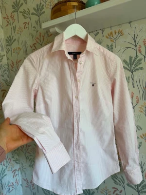  Gant  - Klassisk ljusrosa skjorta från Gant med tunna vita ränder och broderad logga på bröstet. Skjortan har lång ärm, krage och knäppning framtill. Tillverkad i mjuk bomull som känns skön mot huden. Perfekt för en clean och stilren look.