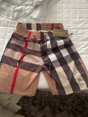 Burberry rutig shorts med snörning - Säljer ett par klassiska Burberry shorts med det ikoniska rutiga mönstret i beige, svart, vitt och rött. Shortsen har snörning i midjan och är tillverkade i ett lätt syntetmaterial. Perfekta för en trendig och avslappnad stil.