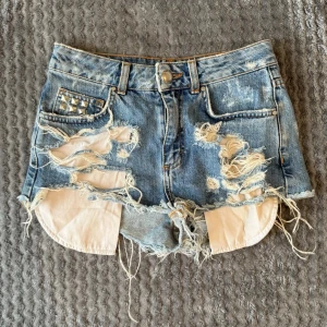 Blå denim shorts från Bik Bok - Säljer ett par jätte snygga blå denim shorts med kraftiga slitningar och råa kanter från Bik Bok. Shortsens ena ficka är dekorerad med silverfärgade nitar. De har låg midja med vita innerfickor som syns tydligt. Älskar dessa med tyvärr så är dom för små. 