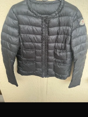 Svart dunjacka från Moncler - Snygg svart dunjacka från Moncler med rund halsringning och fyra framfickor. Jackan har quiltad design, diskret Moncler-logga på ärmen och dragkedja framtill. Tillverkad i glansigt material och fylld med dun för extra värme. Perfekt för kalla vinterdagar. Gammal model, därför ingen qr kod