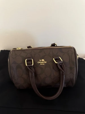 Brun handväska från Coach - Snygg brun handväska från Coach med klassiskt monogrammönster och guldfärgade detaljer. Väskan har två korta handtag och dragkedja upptill. Perfekt storlek för det viktigaste och en stilren accessoar till din outfit.
