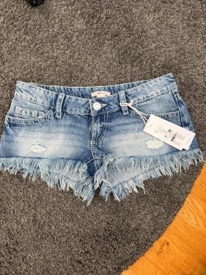 Blå jeansshorts med fransar - Säljer ett par HELT NYA blå jeansshorts från Gina Tricot med slitna detaljer och fransig kant. Lågmidjade! Shortsen har klassisk femficksmodell, bälteshällor och knappgylf. Perfekta för sommaren och ger en avslappnad vibe. Lappen är på, helt nya och aldrig använda. 