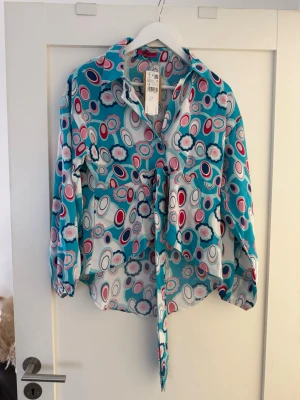 Mönstrad blus med knytband och byxor  - Färgglad blus från Gina Tricot med abstrakt mönster i blått, vitt, rosa och marin. Blusen har lång ärm med resår vid ärmslut, klassisk krage och knytband framtill. Tillverkad i ett lätt och luftigt material som ger en avslappnad look.