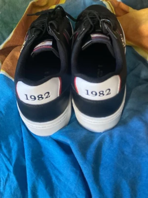 Svarta Beverly Hills Polo Club sneakers - Snygga svarta sneakers från Beverly Hills Polo Club med vita sulor och detaljer i rött, vitt och guld. På hälen står det '1982' och sidan har klubbens ikoniska logga i guld. Skorna har svarta snören och klassisk låg profil. Materialet är syntet med textildetaljer.