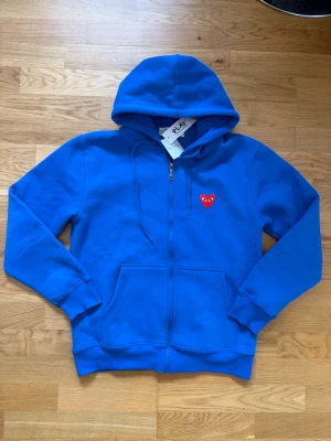 Blå hoodie från Comme des Garçons - Blå zip-hoodie från Comme des Garçons PLAY med röd hjärtlogga på bröstet. Hoodien har dragkedja, två fickor framtill och justerbar huva. Tillverkad i mjuk bomull, perfekt för en chill och stilren look. Står M i tröjan men sitter som S