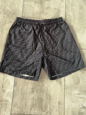 Svarta Fendi shorts med logomönster - Snygga svarta shorts från Fendi med all-over logomönster i grått. Shortsen har snörning i midjan med metalländar och gul detalj. Perfekta för en trendig och avslappnad stil.