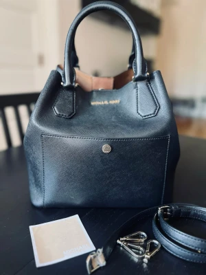Svart handväska från Michael Kors - Snygg svart handväska från Michael Kors i strukturerat läder med guldfärgade detaljer. Väskan har både handtag och avtagbar axelrem samt en praktisk ytterficka med knapp. Insidan är beige. Väskan har några mindre defekter som är lätta att åtgärda, vilket framgår av bilderna. #michaelkors