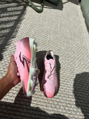 Rosa fotbollsskor från Mizuno - Säljer ett par snygga rosa fotbollsskor från Mizuno med svart logga och coola detaljer. Skorna har en ovandel i syntetmaterial och sulan är skimrande med flera dobbar för bra grepp på planen. Perfekta för dig som vill sticka ut på matchen!