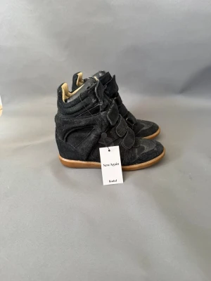 Svarta Isabel Marant wedge sneakers - Svarta sneakers från Isabel Marant med kilklack, gjorda i mocka och textil. Skorna har kardborreband över vristen och en brun gummisula. Hög modell som ger en cool siluett och extra höjd. Perfekta för dig som vill ha en edgy streetstyle-look.