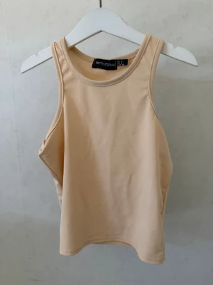Beige crop linnetopp från PrettyLittleThing - Snygg beige linnetopp som är cropped från PrettyLittleThing med hög halsringning och ärmlös design. Toppen har en clean look utan mönster eller detaljer och är gjord i ett mjukt, stretchigt material som sitter bekvämt på kroppen.