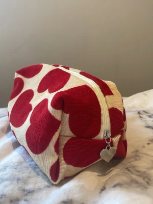 Necessär med röda hjärtan - Söt necessär i tyg med stora röda hjärtan på beige botten. Rektangulär form och dragkedja med hjärtformat metallhänge. Perfekt för smink eller småprylar. Mjuk och rymlig design som sticker ut i väskan.