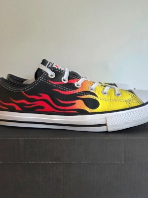 Converse sneakers med flames - Snygga Converse sneakers med lågt skaft, svart bas och coolt flame-mönster i rött, orange och gult. Klassisk vit tå och sula, vita skosnören och detaljer i canvas. Perfekta för dig som vill sticka ut med din stil.