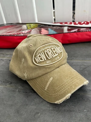 Grön Beige keps med New Orleans-brodyr - Snygg beige keps med broderad text 'NEW ORLEANS' framtill och slitna detaljer för en trendig look. Justerbar baktill och tillverkad i bomullstyg. Perfekt accessoar för en avslappnad streetstyle.