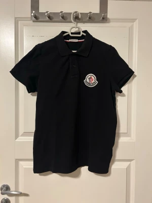 Svart Moncler pikétröja  - Snygg svart pikétröja från Moncler med klassisk krage och knappslå. Tröjan har ett stort Moncler-märke på bröstet och är gjord i mjuk bomull. Perfekt för dig som gillar stilrena och exklusiva plagg.
