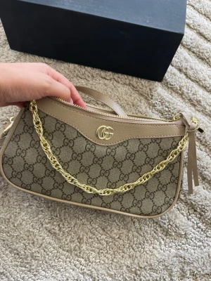 Beige axelremsväska  - Snygg beige handväska  med klassiskt GG-mönster, guldfärgad kedja och justerbar axelrem i läder. Väskan har en stilren, avlång form och detaljer i beige . Perfekt för dig som gillar lyxiga accessoarer.