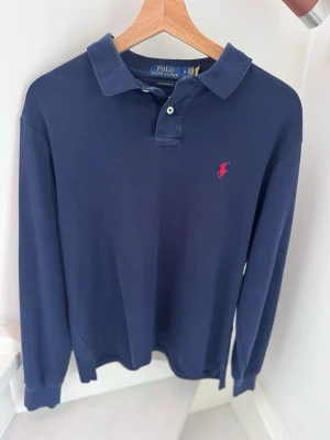 Marinblå långärmad piké från Polo Ralph Lauren - Säljer en blå långärmad pikétröja från Ralph Lauren med marinblå logga. Klassisk modell i Slim Fit. Tröjan är använd och har ett naturligt begagnat skick, men är fortfarande hel och ren. Perfekt för nu inför sommaren  • Märke: Polo Ralph Lauren • Modell: Långärmad Piké • Storlek: Medium men sitter som Small (Slim Fit) • Färg: Blå • Skick: Bra begagnat skick (använd)     •Nypris: ca 1500kr