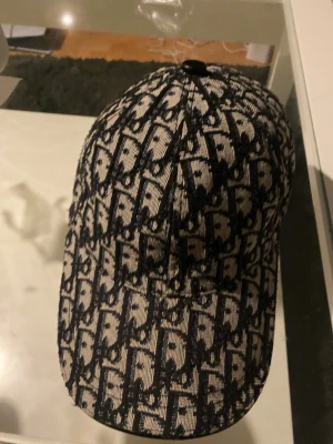  keps från Dior - Säljer en snygg keps från Dior med det klassiska monogrammönstret i svart och beige. Kepsen har böjd skärm och är tillverkad i ett tygmaterial med tydlig struktur. Perfekt för dig som vill ha en statement-accessoar.