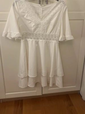 Vit kort klänning med spetsdetaljer - Söt vit kortklänning med v-ringning och vackra spetsdetaljer längs halsringningen och midjan. Klänningen har volang vid ärmslut och kjol, samt korta ärmar. Perfekt för sommarens alla tillfällen.