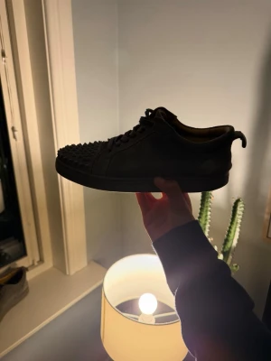 Svarta sneakers med nitar Christian Louboutin - Säljer ett par svarta sneakers från Christian Louboutin med coola nitar över hela tån. Skorna har klassisk låg profil, svart ovandel i läder och den ikoniska röda sulan. Perfekta för dig som vill sticka ut med en edgy och exklusiv look.