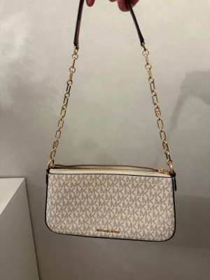 Beige Michael Kors handväska - Snygg beige handväska från Michael Kors med MK-logga över hela väskan. Väskan har guldfärgad kedjerem och dragkedja upptill. Perfekt storlek för det viktigaste och ger en lyxig känsla till din outfit. Aldrig använd och nypris 2700kr