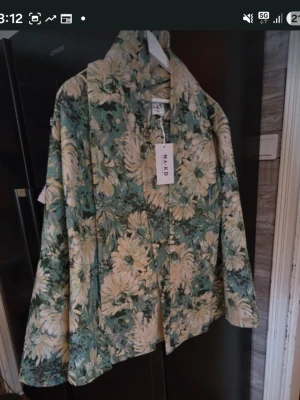 Blommig kappa från NA-KD - Unik kappa från NA-KD med stort blommönster i grönt, beige och vitt. Rak passform, bred krage och knäppning framtill. Perfekt statement-plagg för dig som vill sticka ut med en färgglad och trendig look. Tillverkad i ett mjukt tyg som ger en lyxig känsla. Är oversize i storleken. 