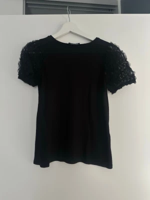 Svart topp med spetsärmar från Zara - Supersnygg svart topp från Zara med korta puffärmar i spets som ger en extra cool touch. Perfekt till jeans eller kjol när du vill ha något lite extra. Klassisk rund halsringning och rak passform.