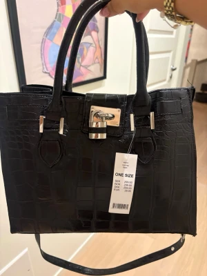 Svart croco handväska med låsdetalj - Stilren svart handväska i croco-mönstrat konstläder med silverfärgade metalldetaljer och lås framtill. Väskan har både dubbla handtag och en justerbar axelrem för flexibel användning. Perfekt för dig som gillar trendiga accessoarer.