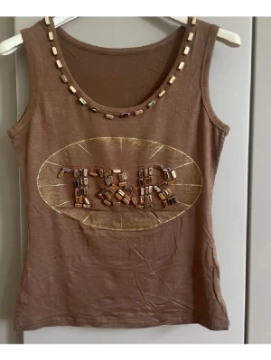 Brunt linne med pärldetaljer - Snyggt brunt linne med rund halsringning och dekorativa pärlor i beige och bruna toner längs halsen och på bröstet. Framsidan har ett ovalt mönster med pärlor som bildar texten 'TRK'. Tillverkat i mjukt bomullsliknande material.