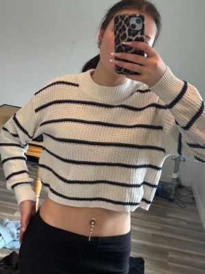 Vit stickad crop tröja med svarta ränder - Säljer en vit stickad crop tröja med svarta horisontella ränder. Tröjan har rund halsringning och ribbade muddar vid ärmslut och nederkant.