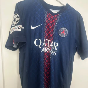 PSG matchtröja Nike blå röd - Snygg PSG matchtröja i mörkblått med röd och vit detalj framtill. Tröjan har Qatar Airways som sponsor, PSG-logga på bröstet och Champions League-märke på ärmen. Tillverkad i lätt och ventilerande material, perfekt för fotboll eller träning. lite skrynklig där bak