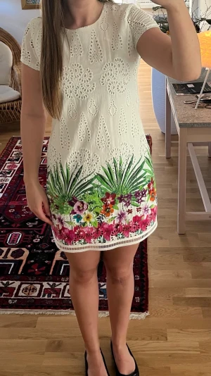 Kortklänning med blommigt mönster - Superfin vit kortklänning med broderade detaljer och blommigt mönster i grönt, rosa och lila längst ner. Klänningen har korta ärmar och rund halsringning. Perfekt för sommarens alla tillfällen!
