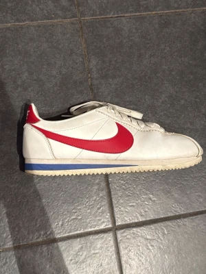 Nike Cortez strl 39 - Klassiska Nike Cortez sneakers i vitt skinn med röd swoosh och blå detalj på sulan. Lågt skaft, vit sula med fiskbensmönster och vita skosnören. Perfekt retrostil för dig som gillar ikoniska sneakers.