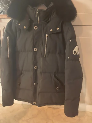 Svart dunjacka Moose Knuckles - Snygg äkta svart dunjacka från Moose Knuckles med klassisk logga på ärmen och flera dragkedjeförsedda fickor. Jackan har stora knappar framtill, quiltad design och hög krage. Perfekt för kalla vinterdagar. 