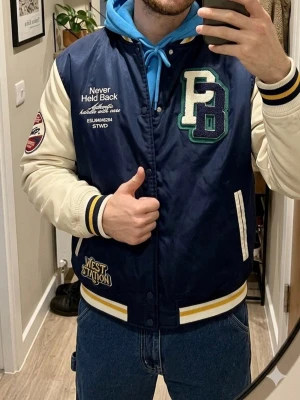 Blå bomberjacka från Pull&Bear - Snygg blå bomberjacka med vita ärmar och broderade patchar, bland annat stora bokstäver på bröstet och textdetaljer. Jackan har ribbade muddar i blått och gult, samt sidofickor. Perfekt för en sportig och trendig look.