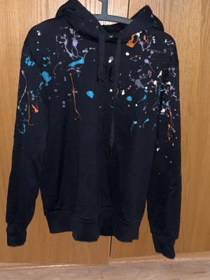 Svart hoodie med färgstänk Dolce&Gabbana - Cool svart hoodie från Dolce&Gabbana med huva och färgstänk i blått, orange, vitt och lila över hela framsidan, ärmarna och huvan. Perfekt för dig som gillar streetwear och vill sticka ut lite extra. Söker hoodien då den inte passar längre. Kvitto från farfetch finns. Ny pris: 10 000! 