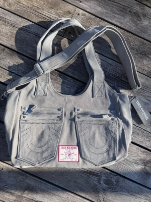 Snygg grå handväska från True Religion🩶 - Snygg grå handväska från True Religion i denim med två framfickor och dekorativa sömmar. Väskan har både handtag och justerbar axelrem samt en tydlig logotyp-patch framtill. 