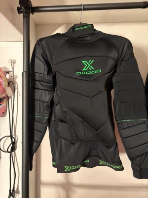 Svart målvaktströja från Oxdog - Tröjan har en grön och svart design samt är tillverkad av högkvalitativa material som skyddar mot hårda skott och snabba spelsituationer. Den är utrustad med SPS (Shoulder Protection System) som förstärker axelområdet och ger extra skydd vid stötar.