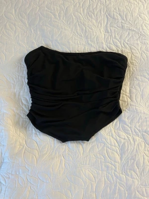 Svart bandeau tubtopp - Säljer en svart bandeau tubtopp med snygga rynkade detaljer framtill. Toppen är axelbandslös och har en croppad passform som passar perfekt till höga jeans eller kjol. Materialet känns mjukt och stretchigt, vilket gör den bekväm att bära hela dagen.