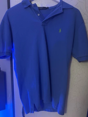 Blå pikétröja från Ralph Lauren - Säljer en klassisk blå pikétröja från Ralph Lauren med korta ärmar och krage. Tröjan har den ikoniska gröna loggan broderad på bröstet och är gjord i bomull för en skön och luftig känsla. Perfekt till jeans eller shorts.