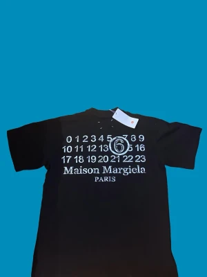Maison Margiela svart t-shirt - Svart t-shirt från Maison Margiela med ikoniskt tryck baktill med siffror och logga. Klassisk passform och rund hals. Tillverkad i mjuk bomull och har korta ärmar. Perfekt för dig som gillar streetwear och vill sticka ut.