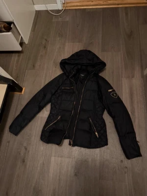 Svart pufferjacka Rockandblue - Snygg svart pufferjacka från Rockandblue med guldfärgade dragkedjor och patch på ärmen. Jackan har huva, flera fickor med dragkedja och quiltade detaljer på sidorna. I storlek S men passar även Xs 