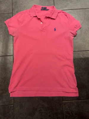 Rosa pikétröja från Ralph Lauren - Säljer en klassisk rosa pikétröja från Ralph Lauren med korta ärmar, krage och två knappar vid halsen. Tröjan har blå broderad logga på bröstet och är tillverkad i mjuk bomull för en bekväm känsla.