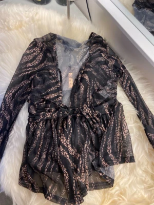 Svart meshblus med mönster från Missguided - Snygg svart meshblus från Missguided med beige mönster och omlott-design. Blusen har v-ringning, långa ärmar och knytband i midjan. Materialet är genomskinligt och ger en trendig look.